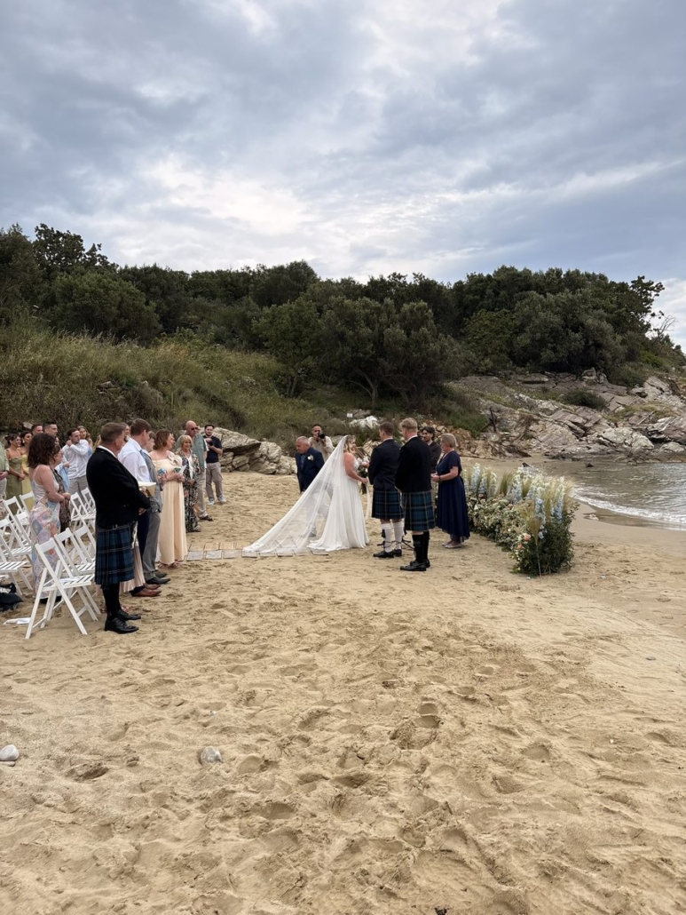 Bride and groom exchanging vows on Skoutari beach, Mani, with guests in kilts and floral arch